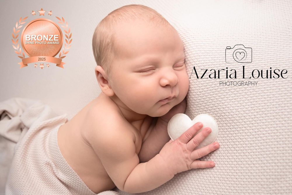 A peaceful sleeping newborn baby rests on soft white fabric while gently holding their tiny hand against their face.