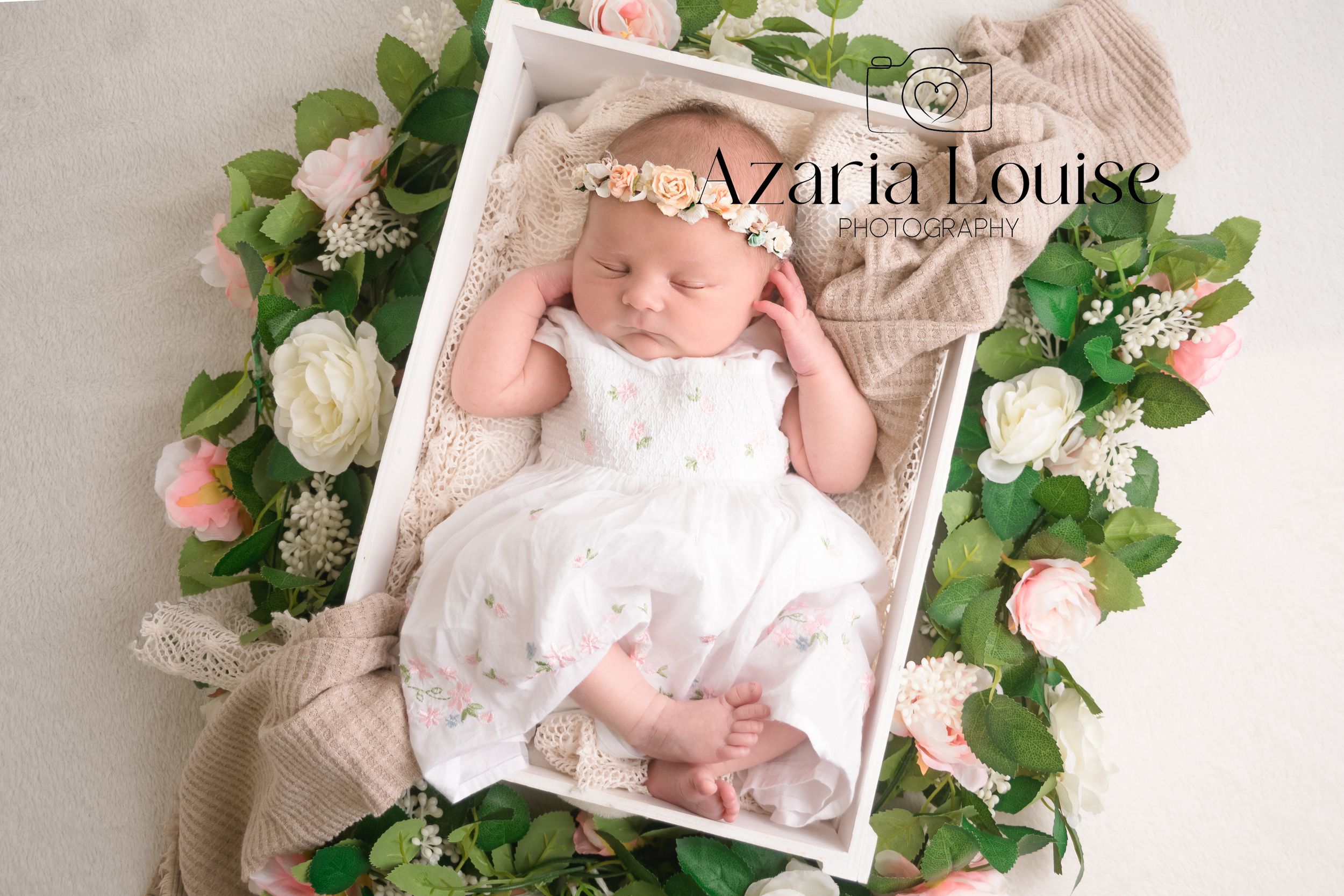 Newborn wearing white dress rests in wooden tray surrounded by pink roses and green foliage on beige backdrop.