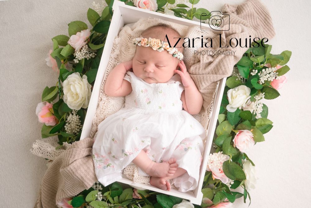 Newborn wearing white dress rests in wooden tray surrounded by pink roses and green foliage on beige backdrop.