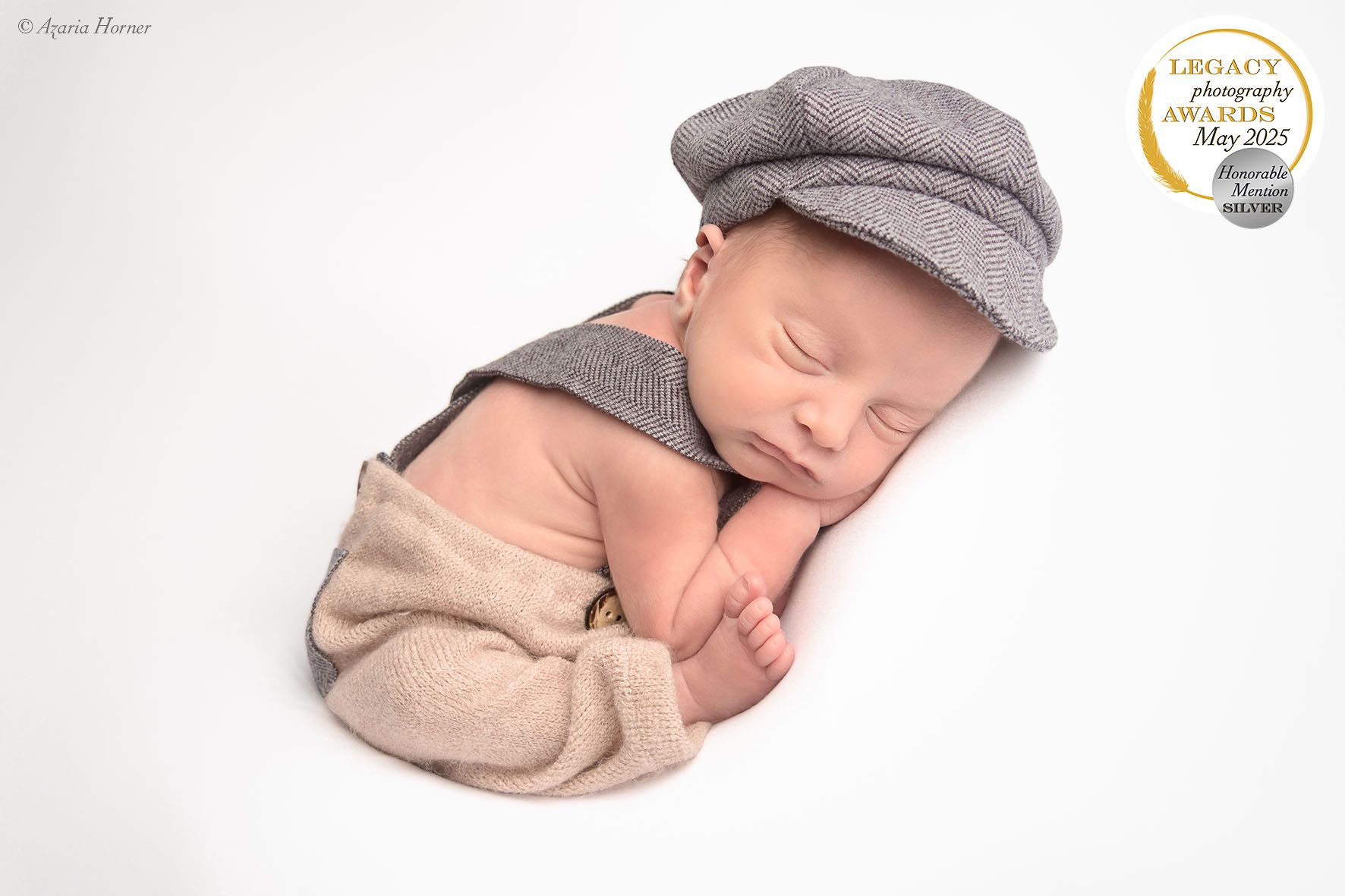 Sleeping newborn wearing gray newsboy cap and beige wrap curled up on white background during professional photo session.