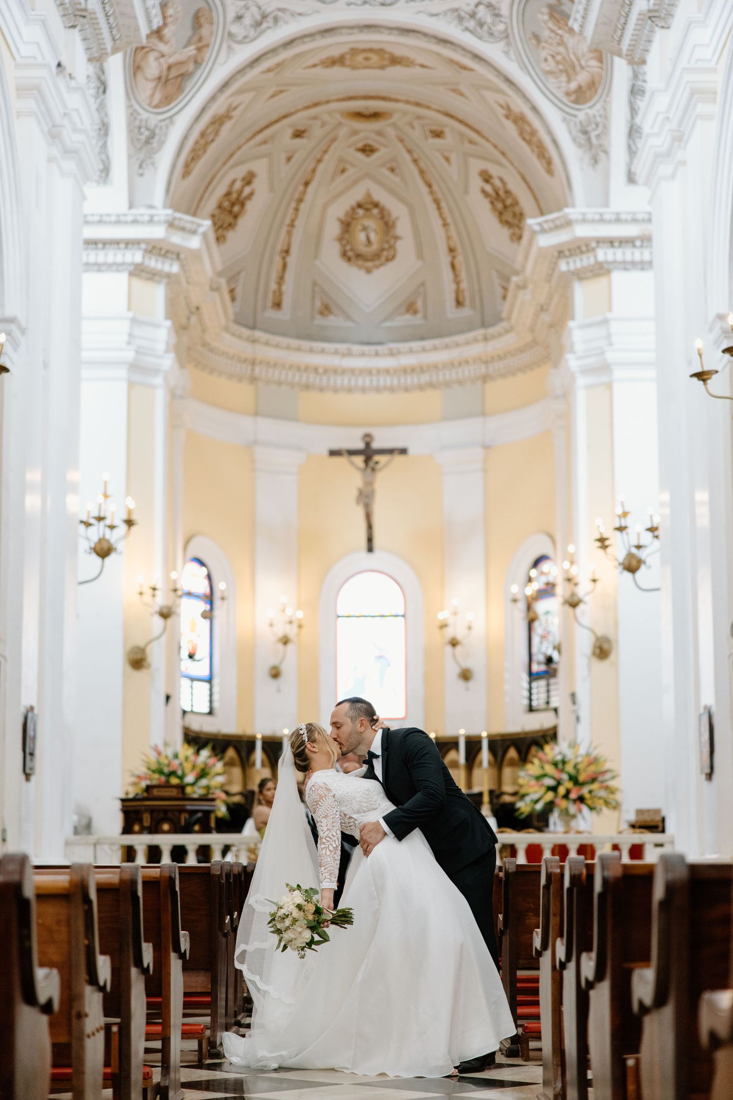 Romantic wedding kiss in ornate white church with high vaulted ceiling.
