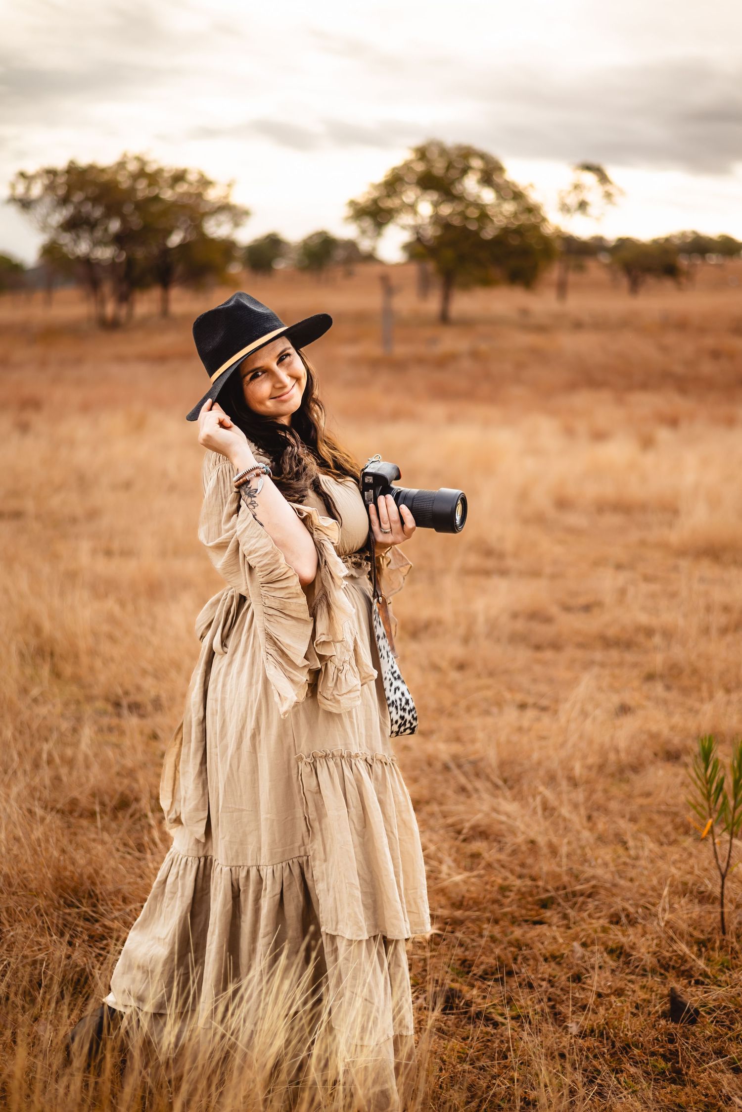 Photographer wearing flowing dress and wide-brimmed hat poses with camera in rural field.