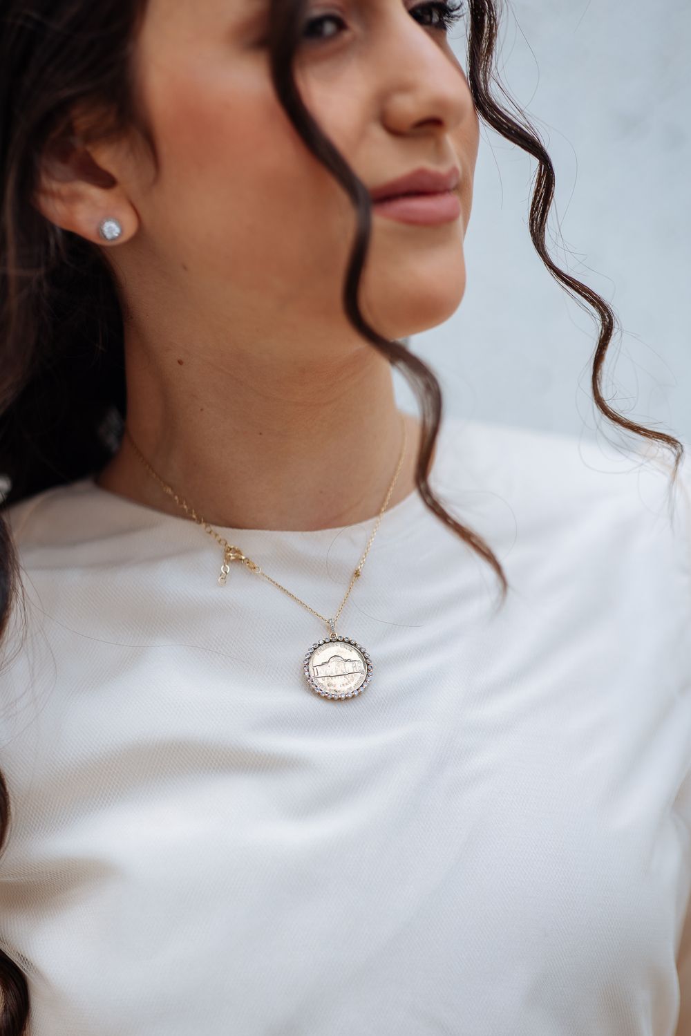 Close-up details of gold chain necklace with round pendant worn with white top against light background.