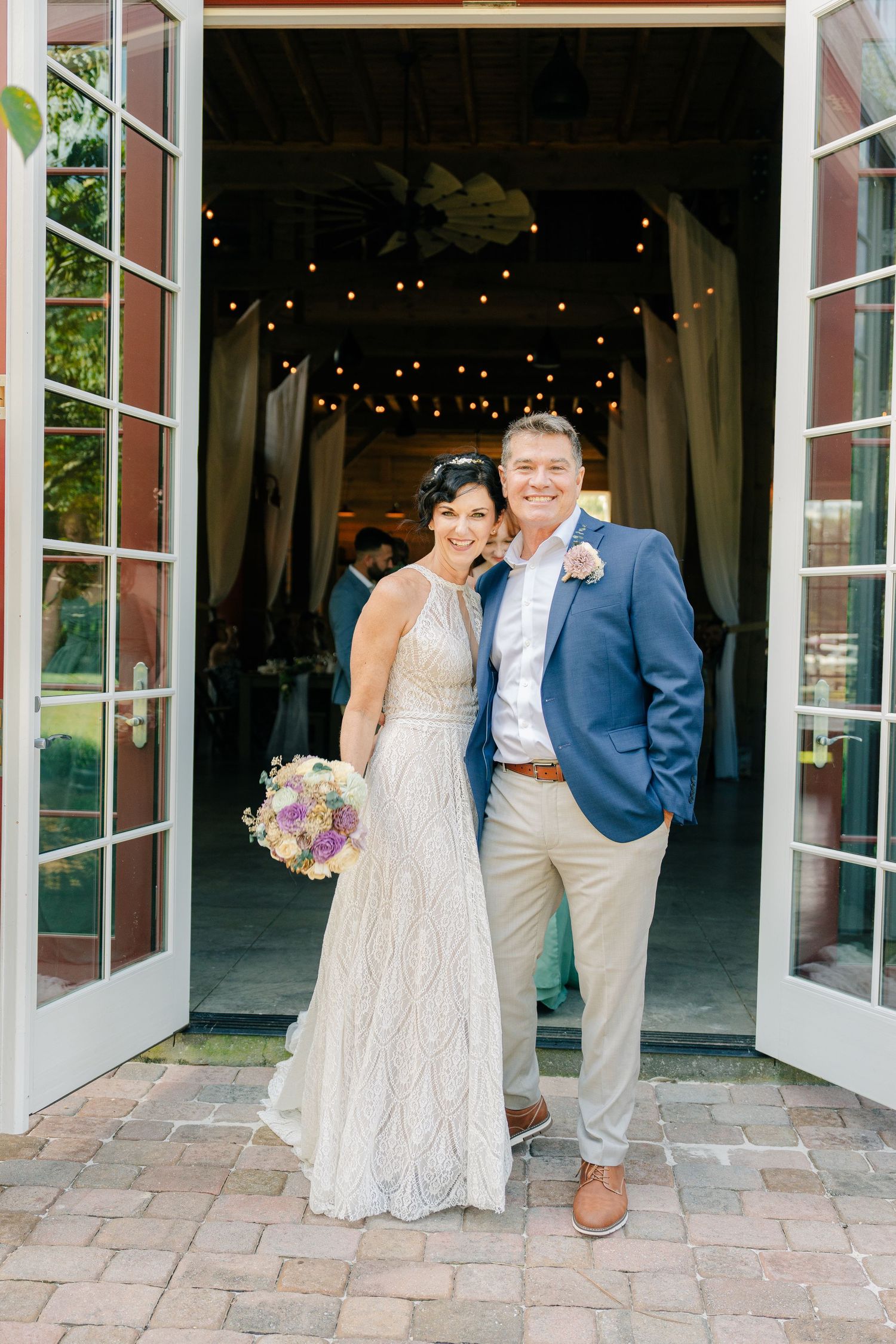 A couple stands in a rustic doorway decorated with string lights while dressed in wedding attire on their special day.