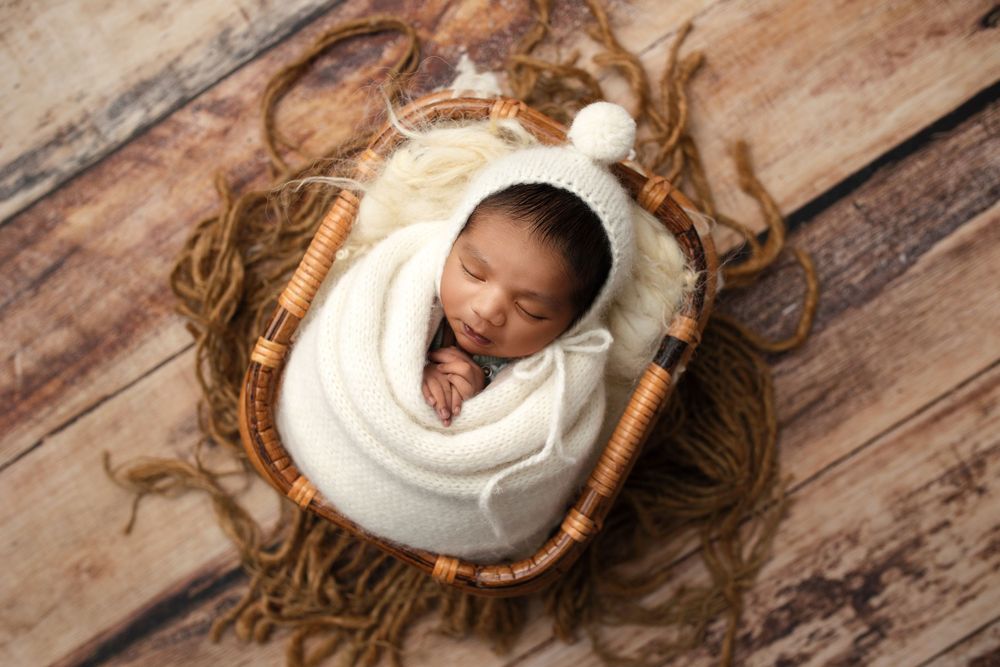 Newborn baby wrapped in white swaddle sleeping peacefully in woven basket on wooden floor.