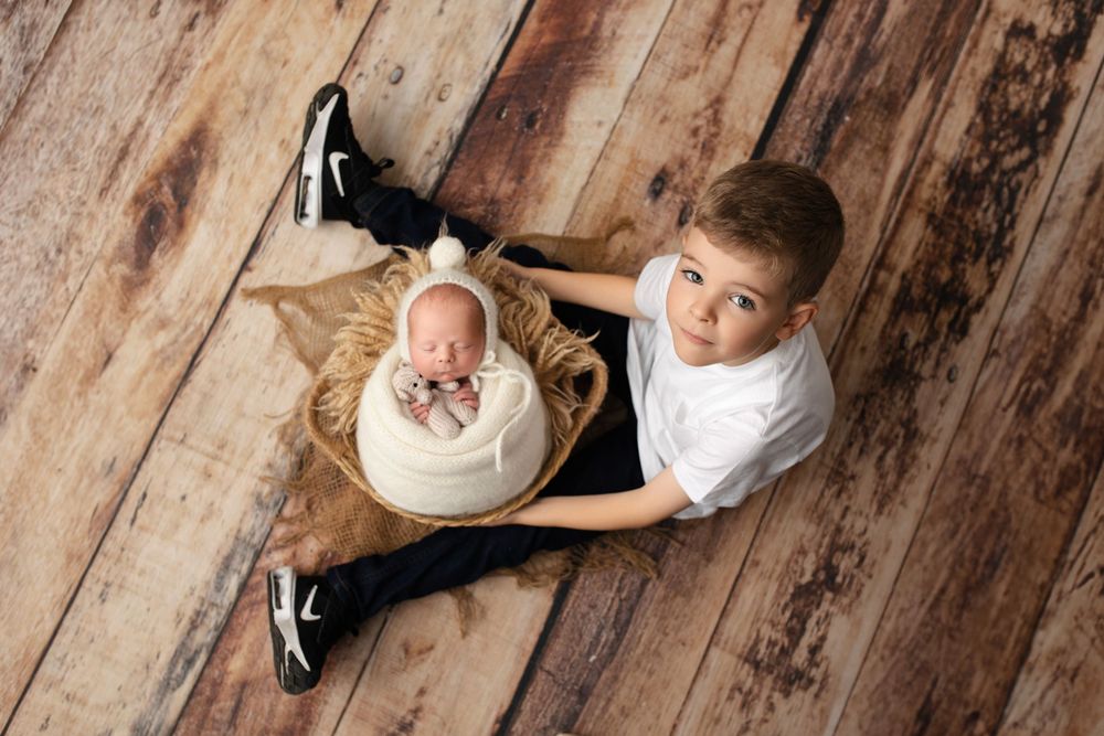 Sweet sibling portrait with baby in basket and older child on wooden floor.
