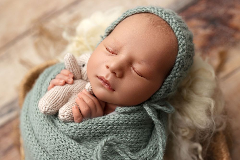 Peaceful sleeping newborn wearing knit teal bonnet and wrap against fuzzy white backdrop.