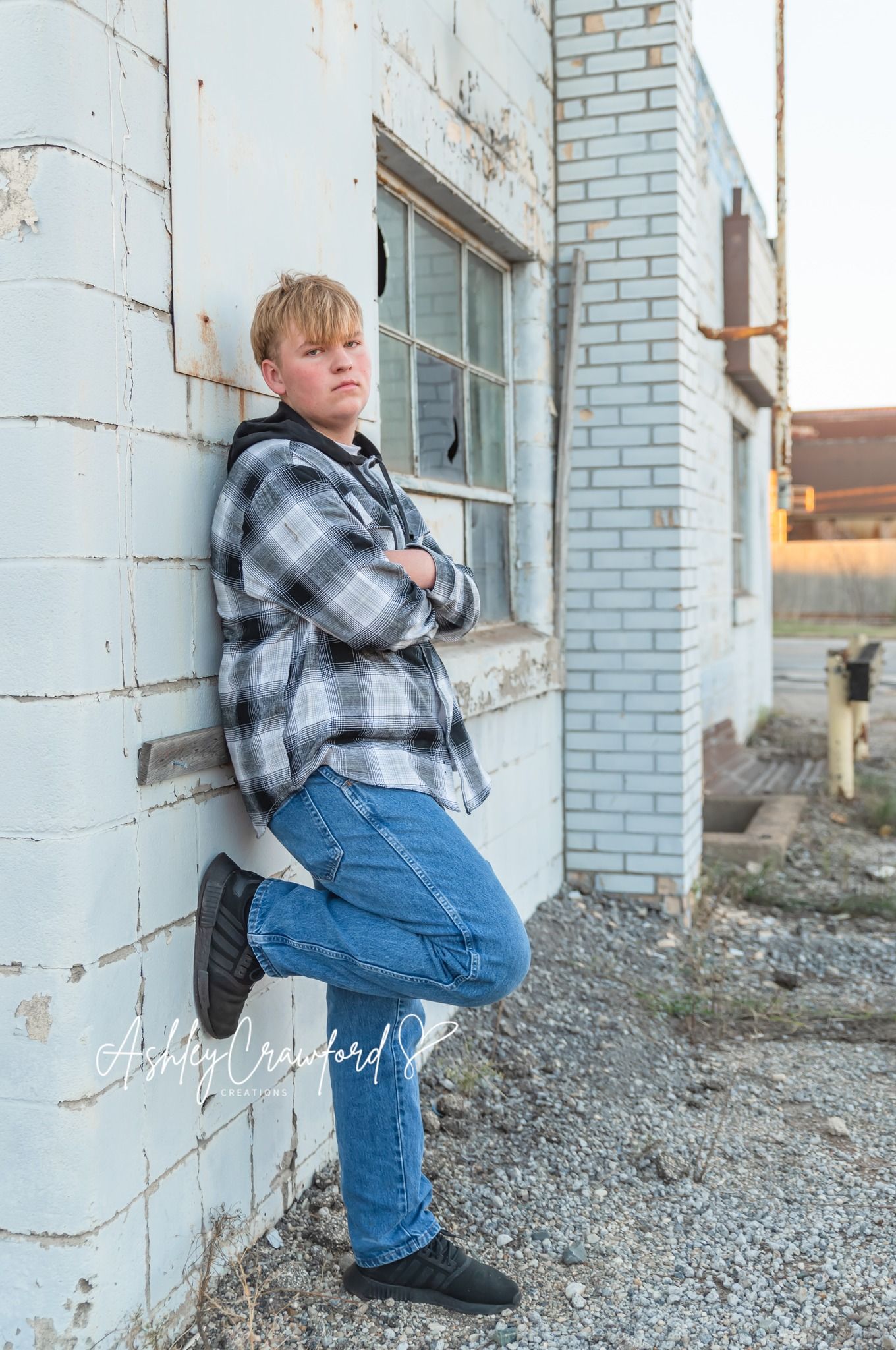 Teen in plaid flannel and jeans leaning against white brick building in casual pose.