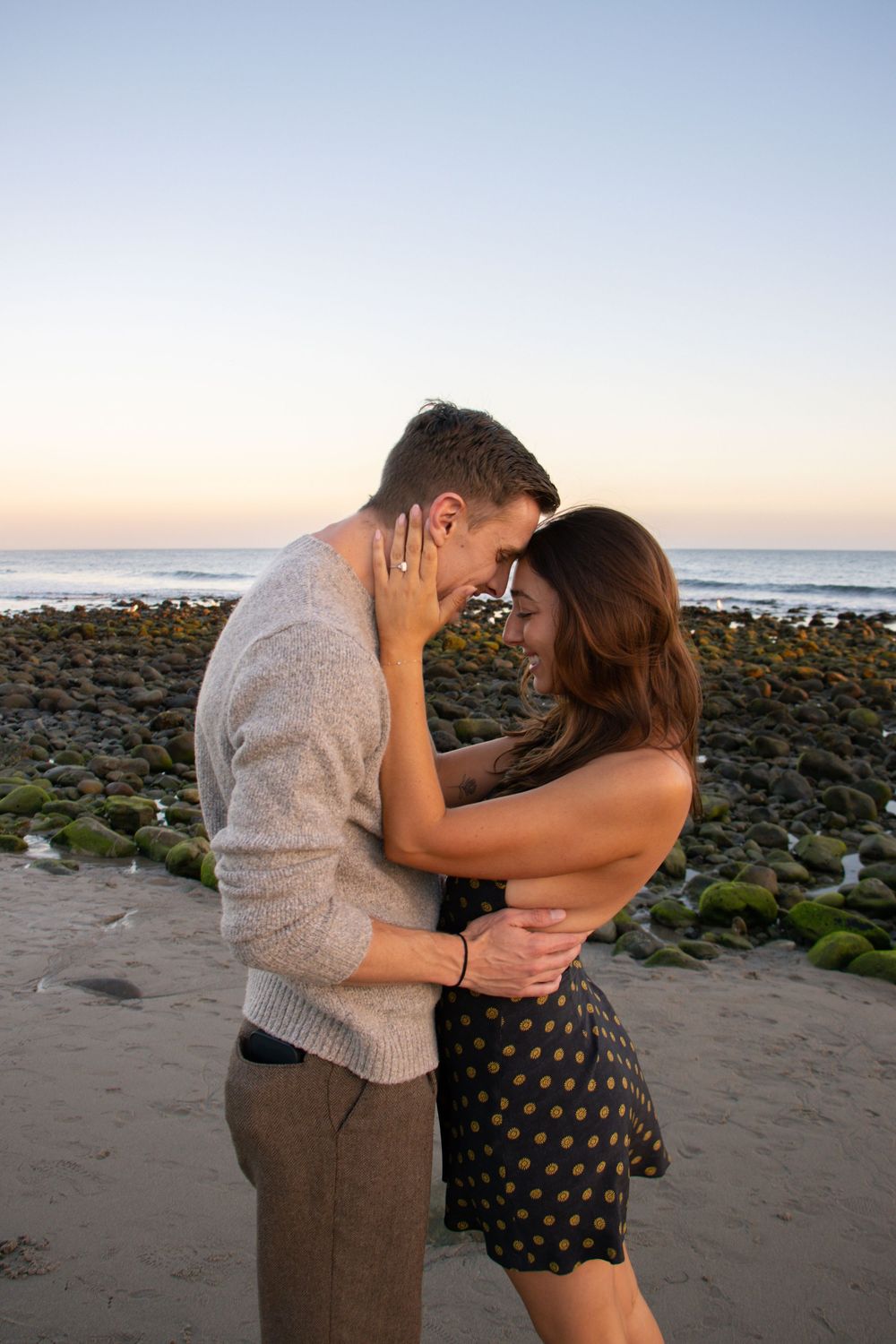 Intimate moment between couple embracing on beach with rocky coastline at dusk.