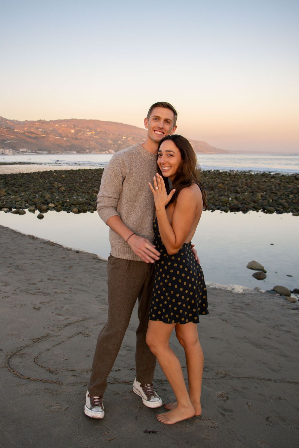 Happy couple posing together on beach with mountains and ocean in background at sunset.