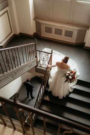 Aerial view of bride in white gown holding bouquet descending grand staircase with groom below at elegant wedding venue.