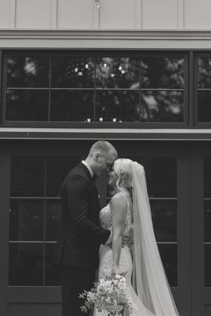 Black and white photo of bride and groom sharing an intimate moment, touching foreheads outside a venue.