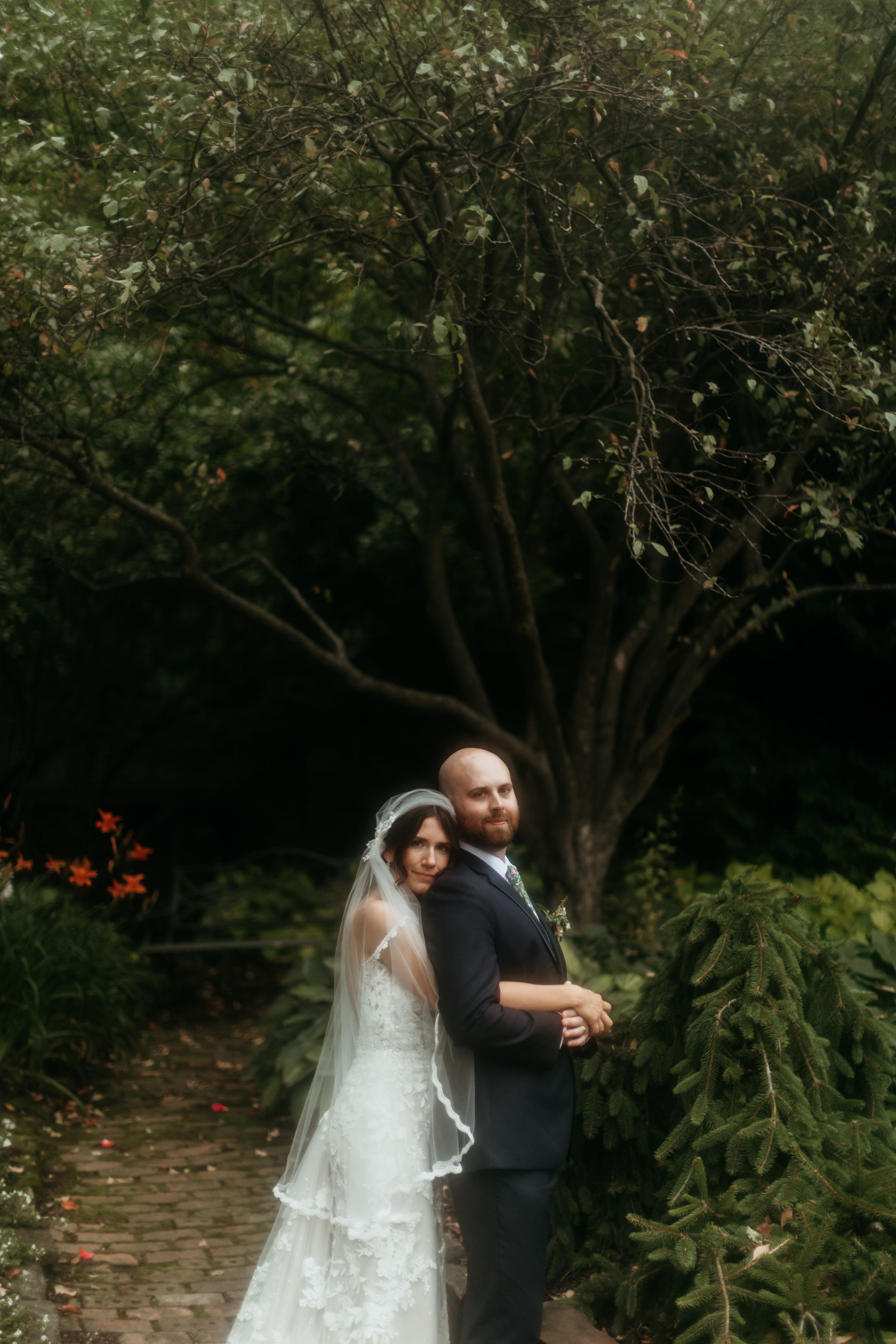 Bride and groom embrace on a garden path surrounded by lush green trees in a romantic wedding portrait.