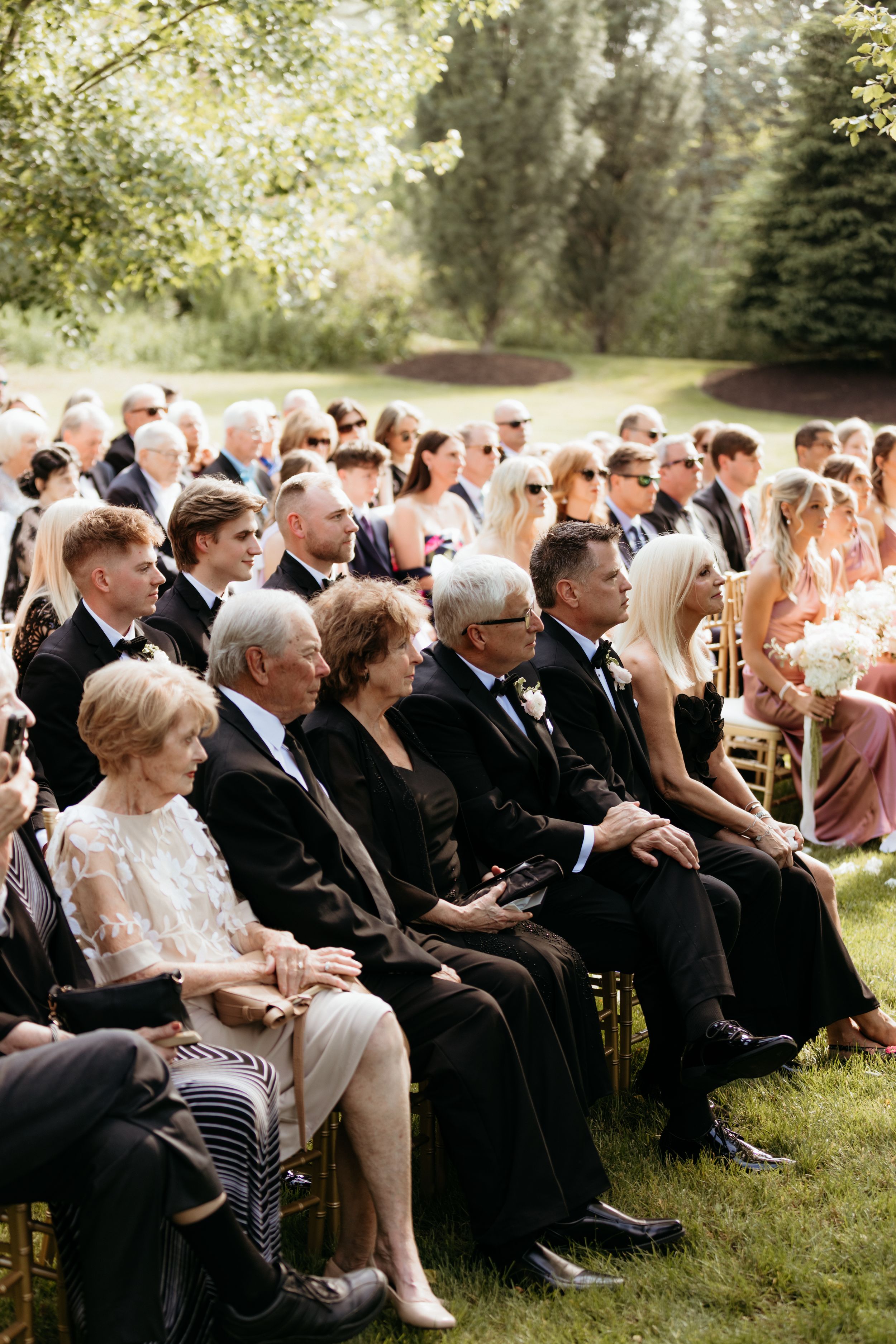 Guests seated in gold chairs at an outdoor wedding ceremony surrounded by lush green trees on a sunny day.