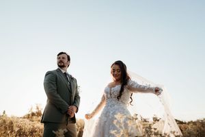 Bride twirls veil in golden field while groom stands beside her in sage suit.