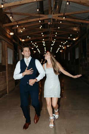 Joyful bride and groom walking hand-in-hand through a rustic barn venue decorated with string lights.