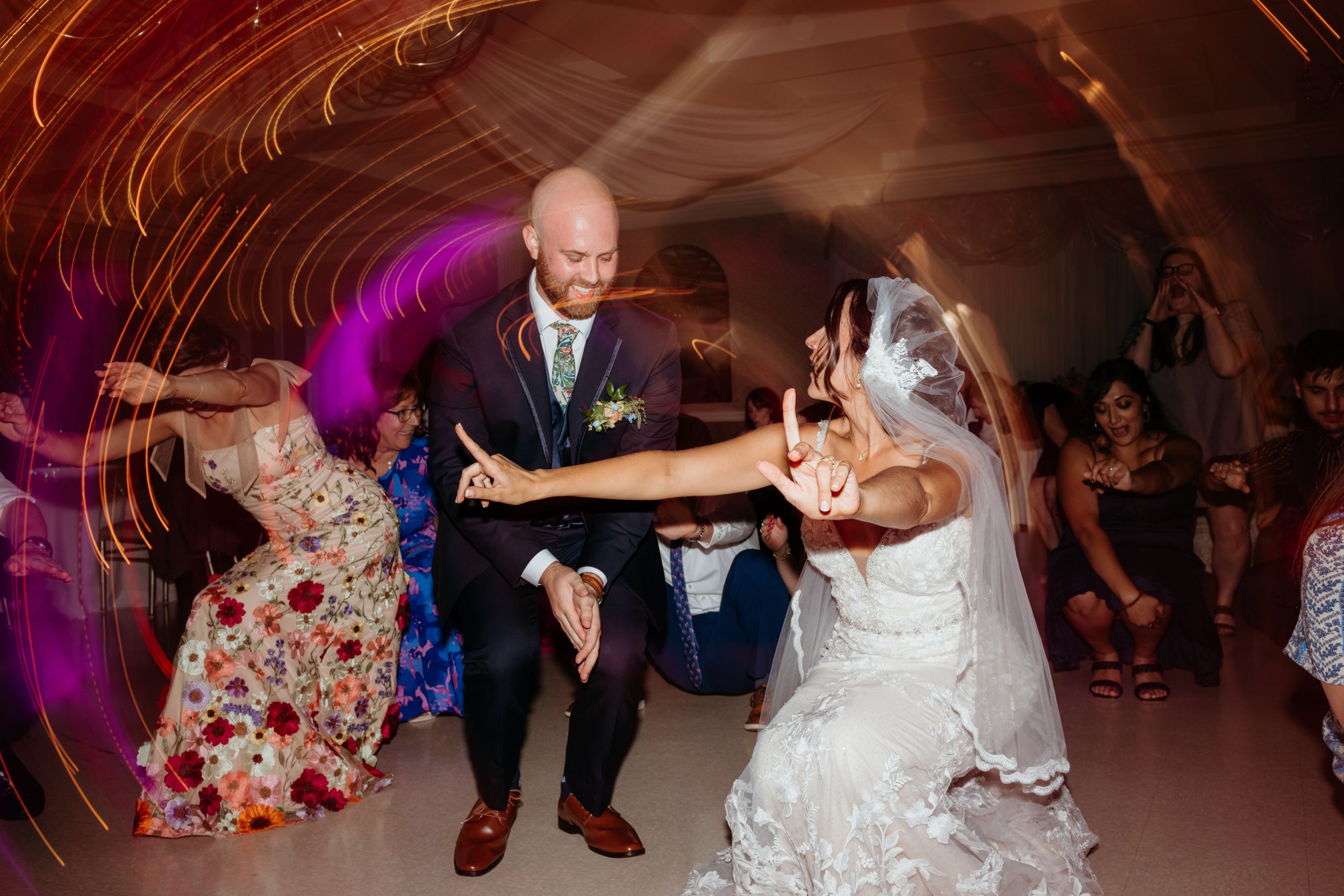 Bride and groom dancing joyfully at wedding reception with colorful light trails and guests watching in background.