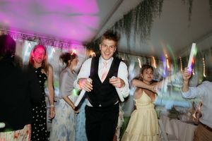 Young man in vest dancing joyfully at a wedding reception party with guests holding glow sticks.