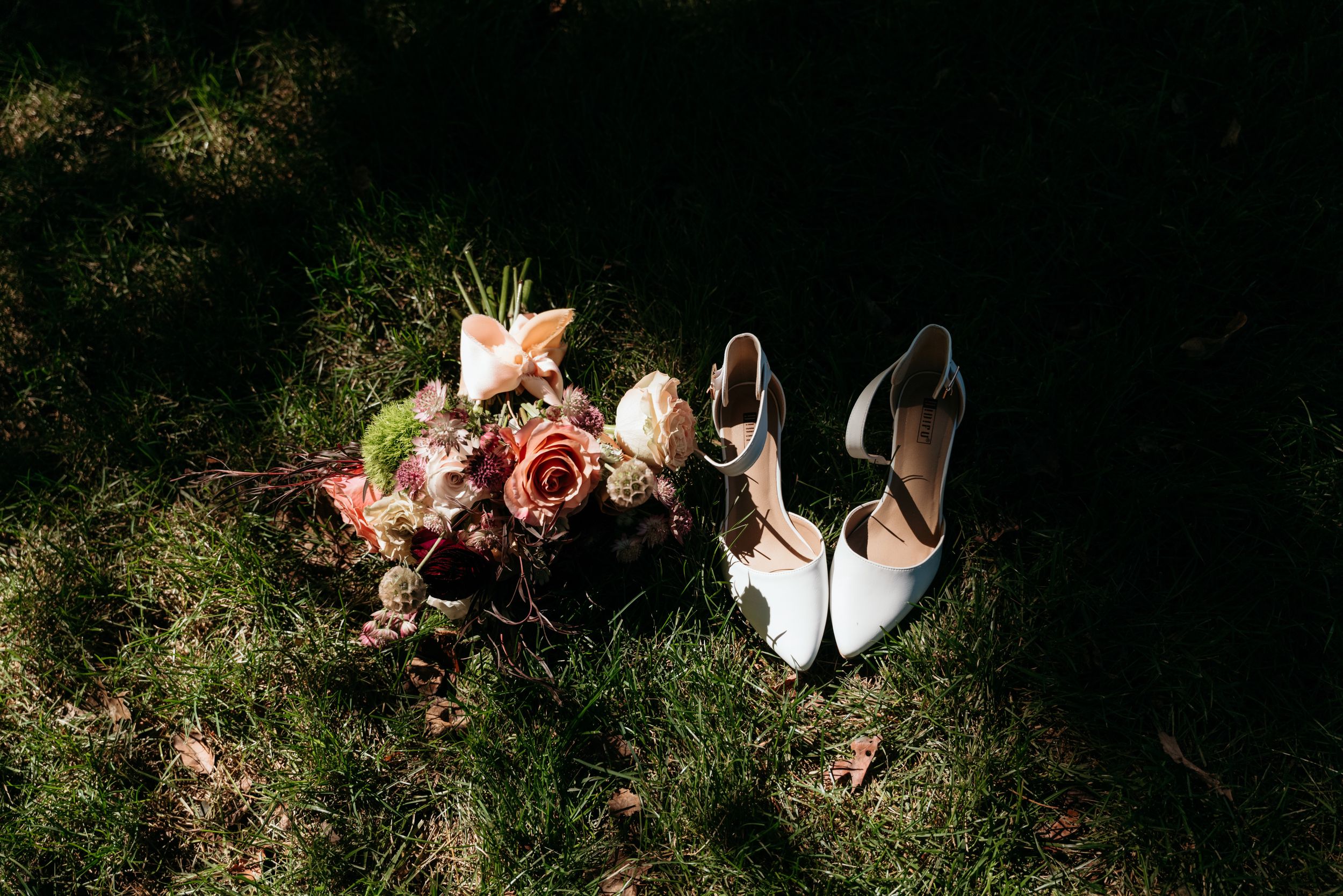 Bridal bouquet of roses and white heels rest on grass in a sunlit wedding detail flat lay.