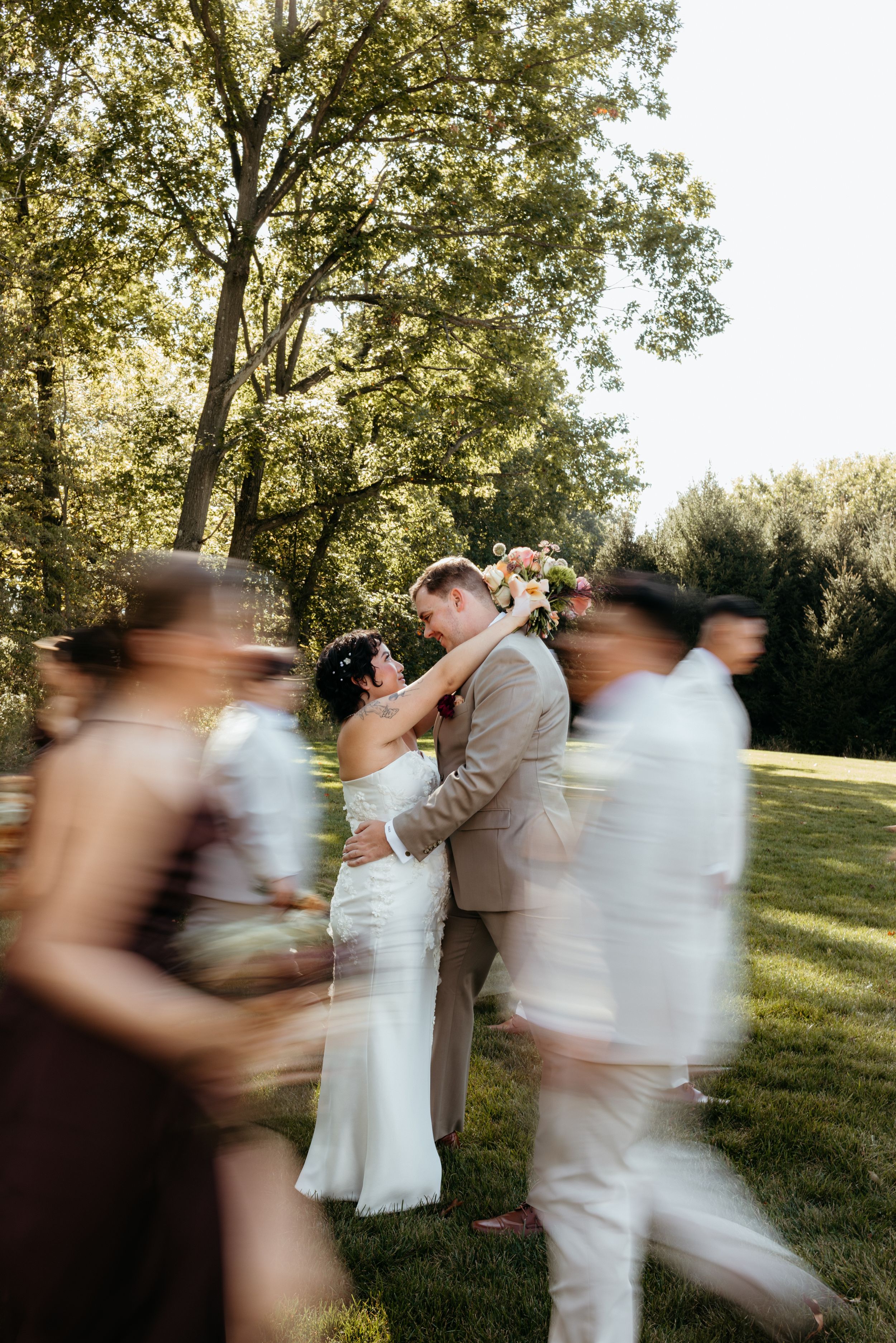 Bride and groom kiss while wedding party walks past in a creative motion-blur outdoor portrait.