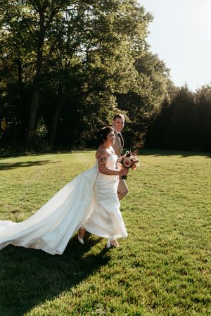 Bride and groom walk joyfully across sunny green lawn, bride holding bouquet with long dress train.