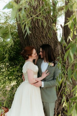 Bride and groom share a romantic moment under a willow tree, bride in white gown, groom in sage green suit.