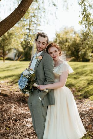 A bride and groom pose together outdoors, she in a cream tulle dress, he in a gray suit holding a blue hydrangea bouquet.