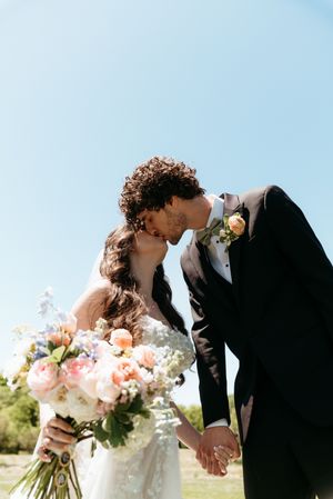 Bride and groom kiss while holding hands outdoors under a bright blue sky.