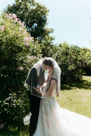 Couple shares a kiss under a flowing veil near blooming lilac bushes outdoors.