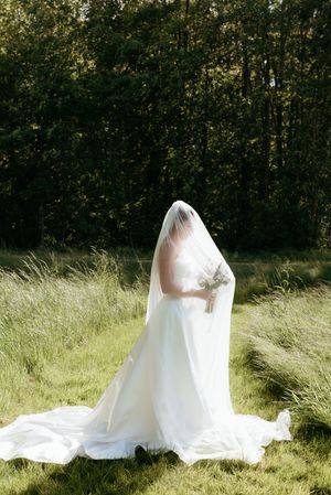 Bride in white gown and veil standing in a sunlit meadow with her face covered by the flowing veil.