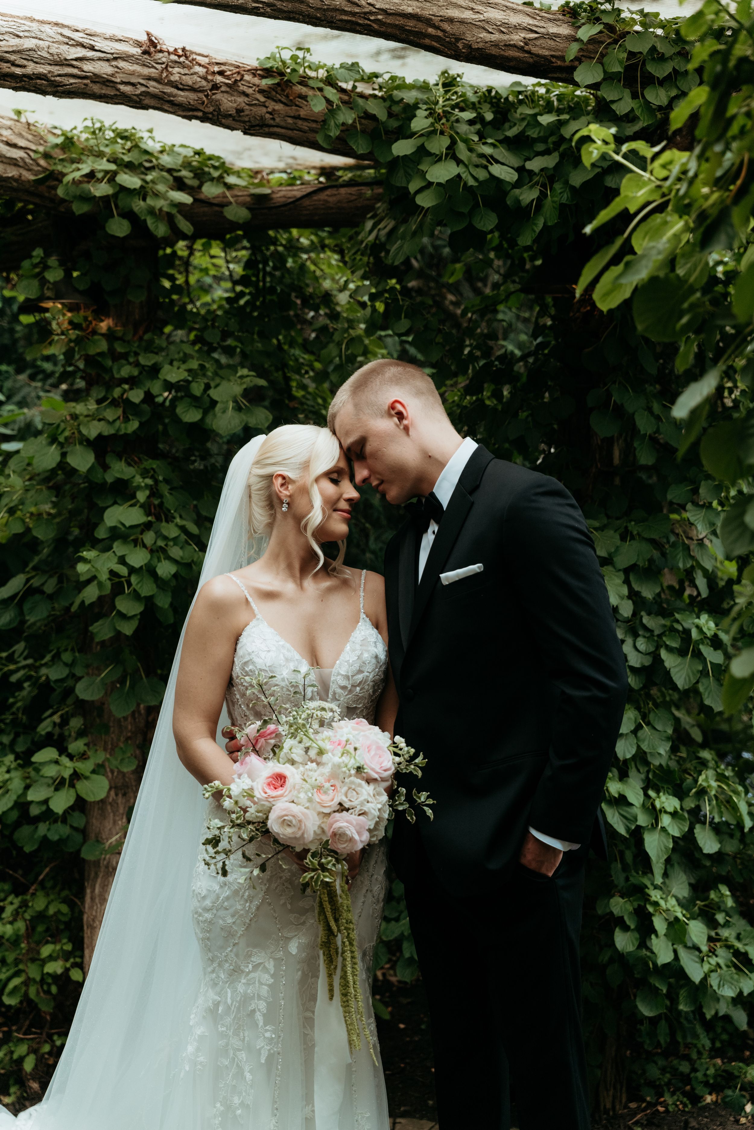 Bride and groom touching foreheads tenderly under rustic wooden arch with lush green ivy backdrop.