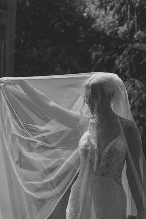 Black and white photo of a bride lifting her flowing veil outdoors on her wedding day.