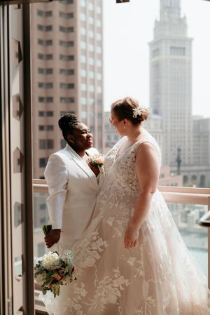 Two brides share a tender moment on a balcony with a city skyline backdrop on their wedding day.
