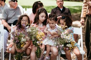 Four young flower girls sitting together holding vibrant wildflower bouquets at an outdoor wedding.