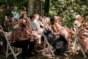 Wedding guests seated on white chairs in a wooded outdoor ceremony setting.