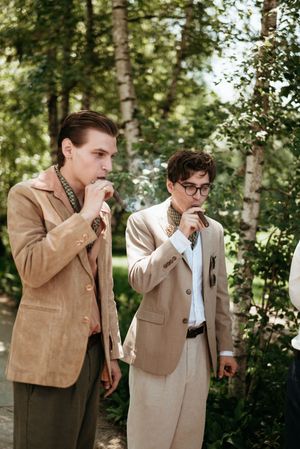 Two groomsmen in beige suits standing outdoors among birch trees at a wedding ceremony.