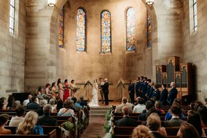 Wedding ceremony inside a stone chapel with stained glass windows, wooden pews, and wedding party at altar.