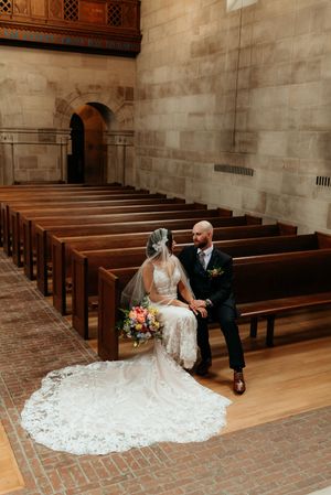 Bride in lace gown and groom sit together on church pews inside a stunning stone chapel.