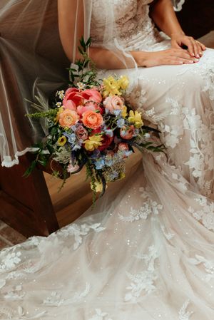 Bride in lace wedding gown seated with colorful spring bouquet of pink, yellow, and blue flowers resting on wooden bench.
