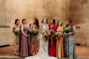 Bride with bridesmaids in colorful mismatched dresses holding bouquets in a stone chapel.