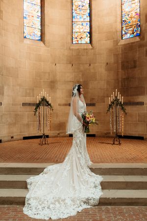 Bride in lace gown with long train stands at chapel altar with stained glass windows.