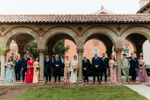 Wedding party in colorful attire poses under arched colonnade of historic building.