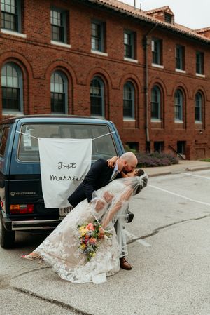 Newlywed couple sharing a romantic dip kiss in front of a Just Married Volkswagen van outside a brick building.