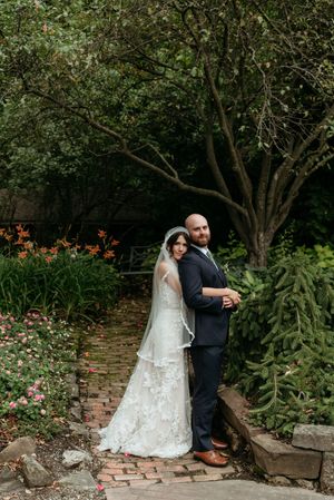 Bride and groom embrace back-to-back on a brick garden path surrounded by lush greenery.