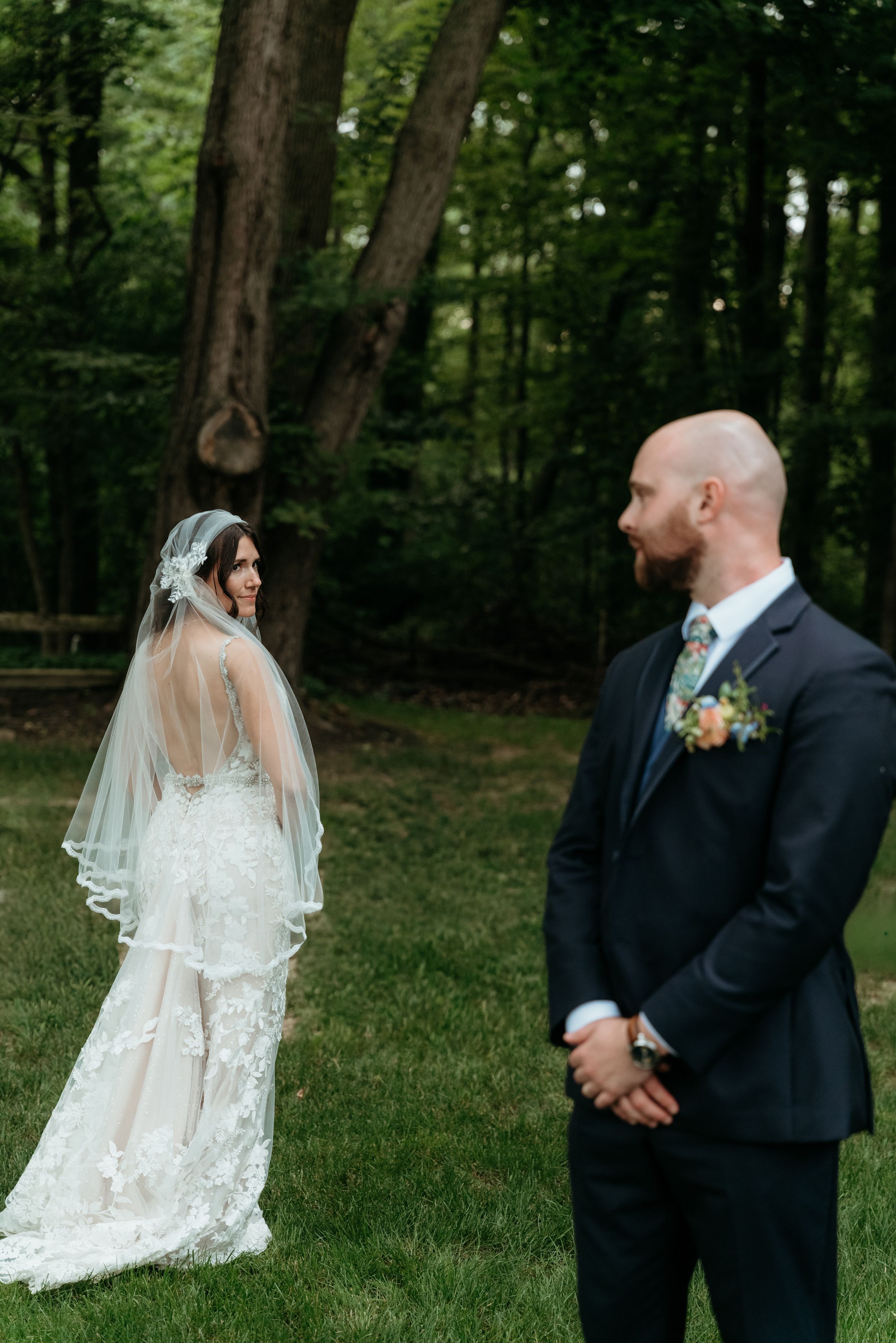 Bride in lace gown and veil looks back at bald groom in navy suit during first look in forest.