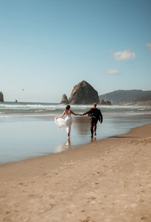 Bride and groom run hand-in-hand along sandy beach with Haystack Rock in background.