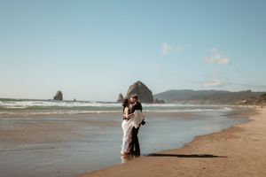 Couple embraces on a sunlit beach with Haystack Rock visible in the background.