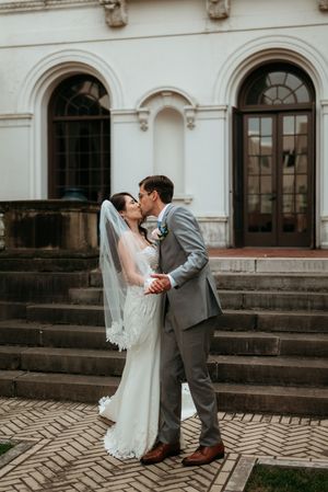 Bride and groom share a romantic kiss on brick steps outside a white classical building, bride wearing lace gown and veil.