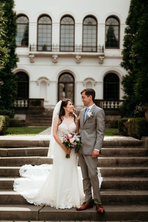 Bride and groom stand on stone steps before elegant white mansion, bride holding colorful bouquet in lace wedding gown.