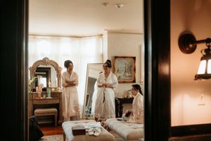 Bridesmaids in white robes preparing for a wedding in an elegant room with a vintage mirror and warm lighting.